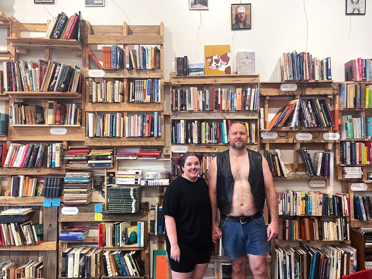 Two people stand in front of a very busy wall of bookshelves. The man on the right is taller, wearing an open black vest with no shirt and denim shorts. The woman on the left is shorter, wearing all black and light cat makeup