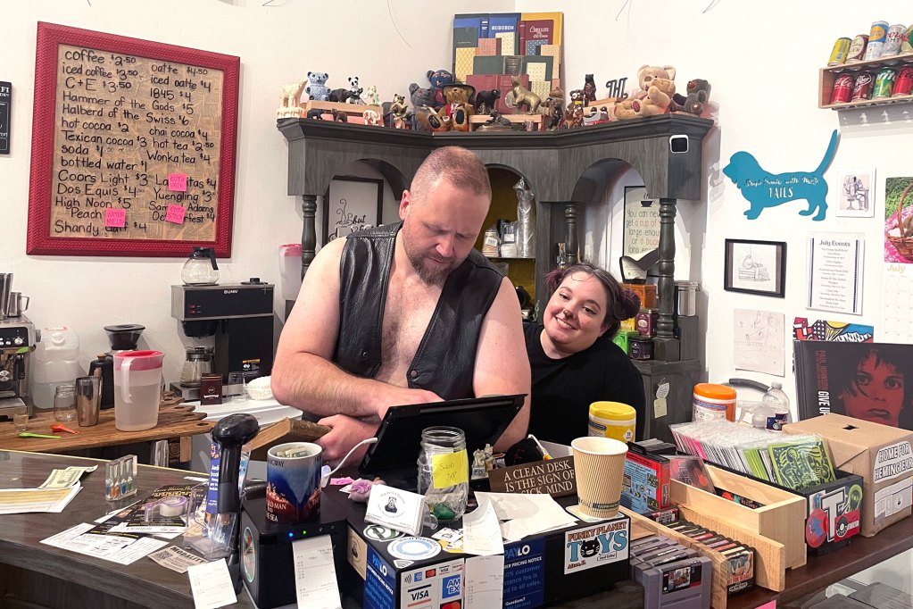 A couple sit behind a busy countertop with items to purchase and a tablet that they look at. Behind them are more decorations and a beverage price board
