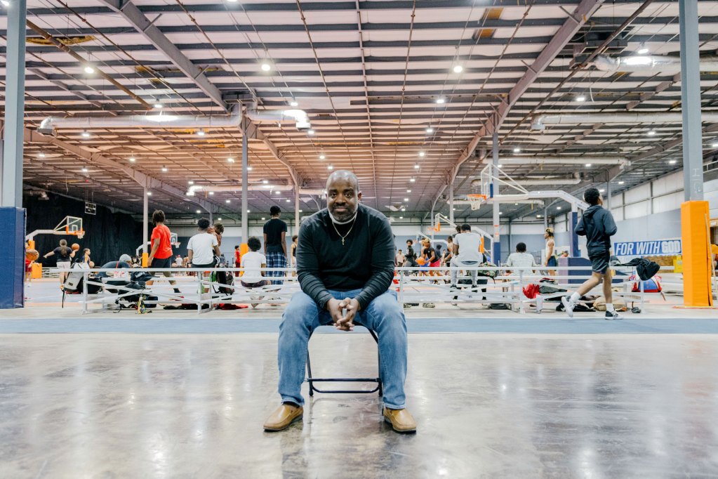 A man in a inside a workout facility poses in a folding chair with hands clasped before him
