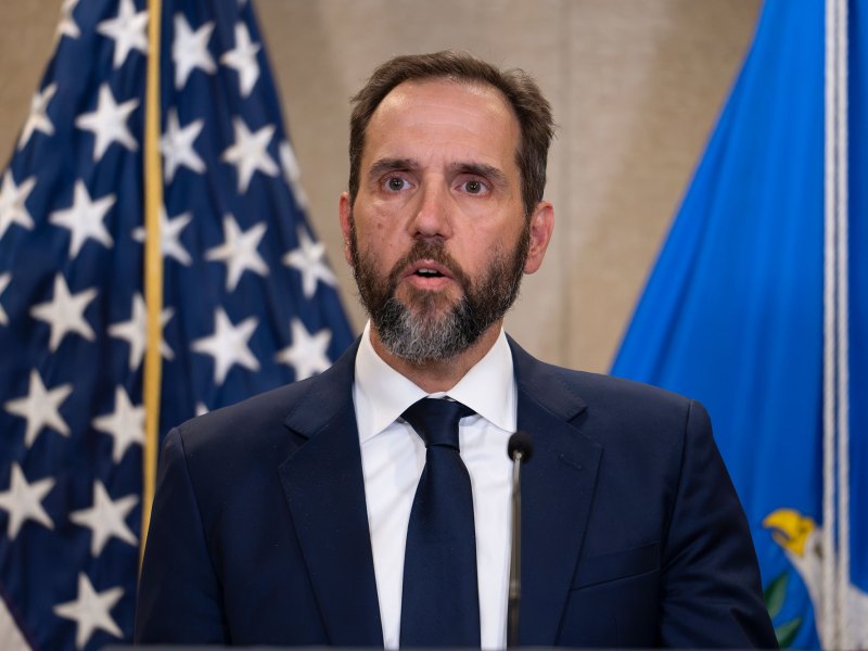 A bearded man in a suit speaks in front of a US flag