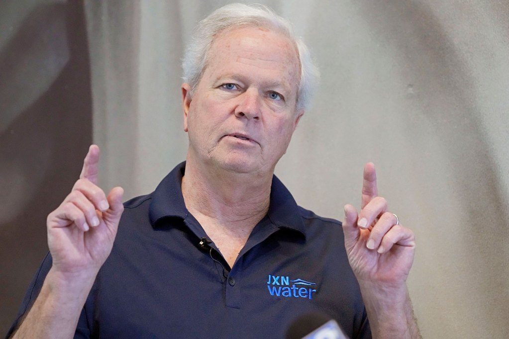 A man wearing a dark polo shirt marked "JXN Water" speaks with two fingers pointing in the air