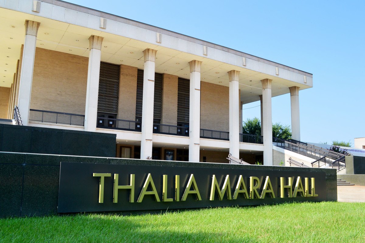 An exterior view of a two story building with tall white columns and a black and gold sign up front that reads Thalia Mara Hall