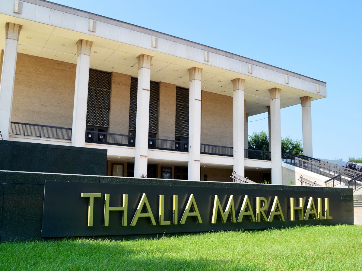 An exterior view of a two story building with tall white columns and a black and gold sign up front that reads Thalia Mara Hall