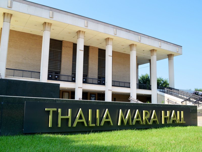 An exterior view of a two story building with tall white columns and a black and gold sign up front that reads Thalia Mara Hall