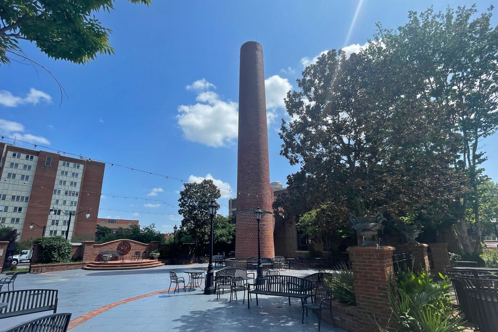 A courtyard lined with a few trees on each side with metal benches for seating, and brick fence details. A large brick chimney stack looms in the background
