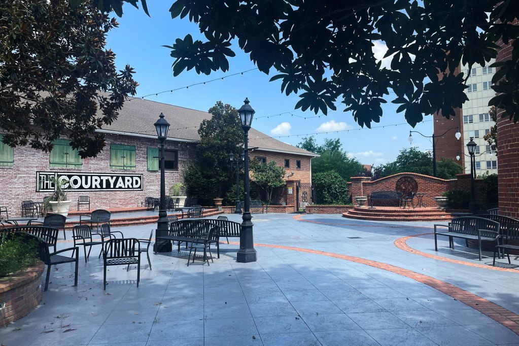 A courtyard lined with a few trees on each side with metal benches for seating, and brick fence details.