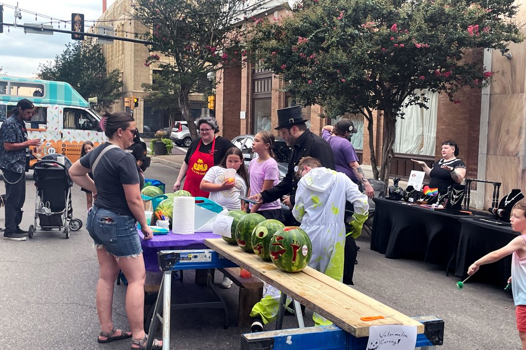 Vendor tables set up in the middle of a street with people walking around them. One in the center displays round watermelons carved like jack o' lanterns