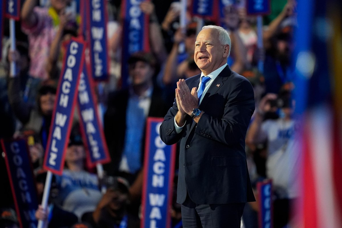 A man in a black suit and blue tie holds his hands together as he speaks to a large crowd. "Coach Walz" signs are visible behind him