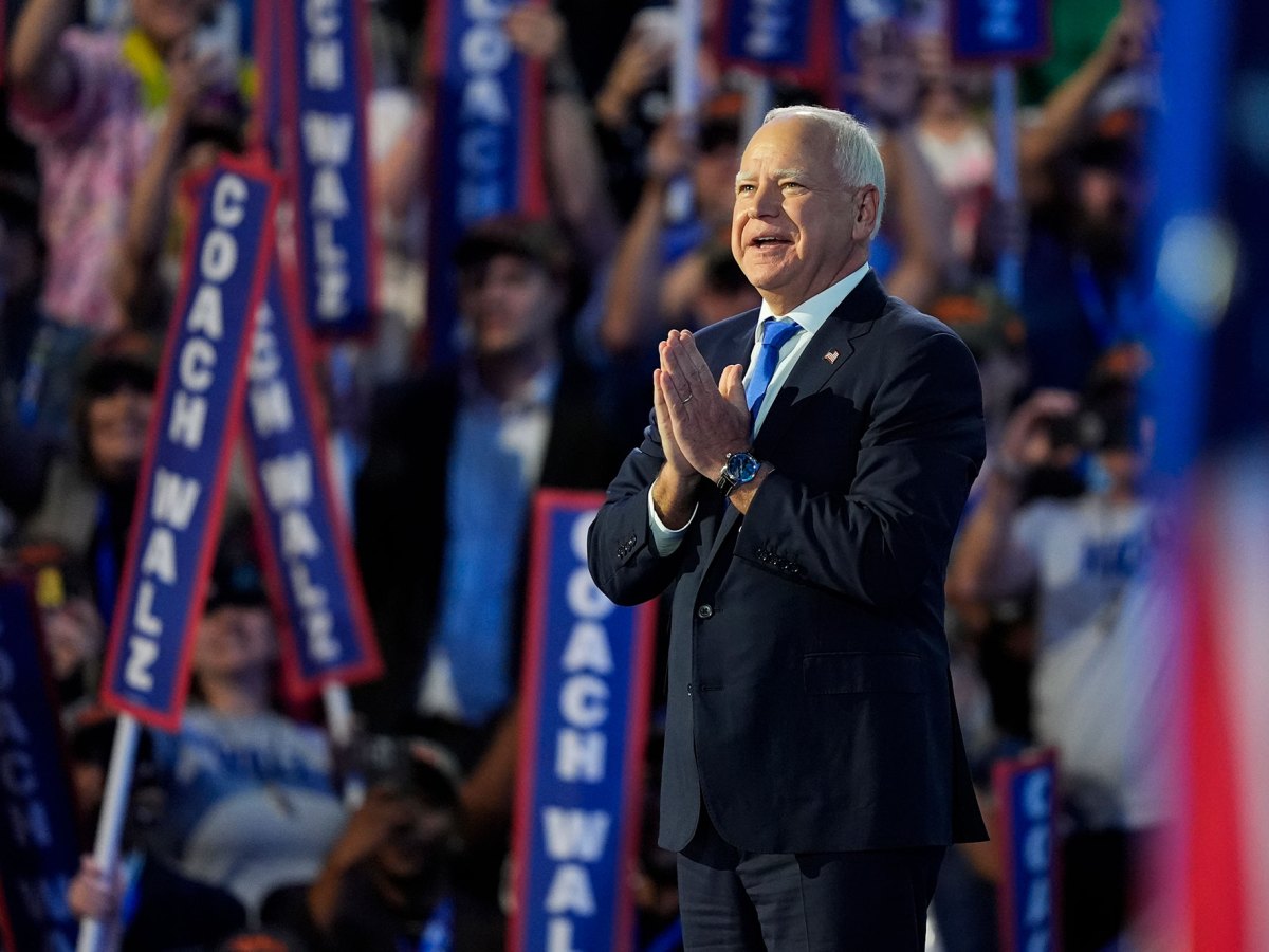 A man in a black suit and blue tie holds his hands together as he speaks to a large crowd. "Coach Walz" signs are visible behind him