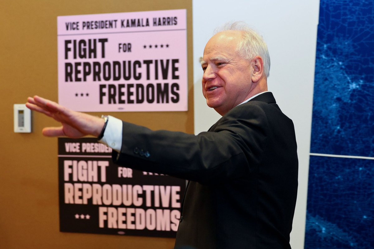 A man in a suit smiles and raises his arm towards the left of the photo. Behind him are signs that read: Vice President Kamala Harris. Fight for Reproductive Freedom