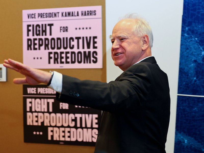 A man in a suit smiles and raises his arm towards the left of the photo. Behind him are signs that read: Vice President Kamala Harris. Fight for Reproductive Freedom
