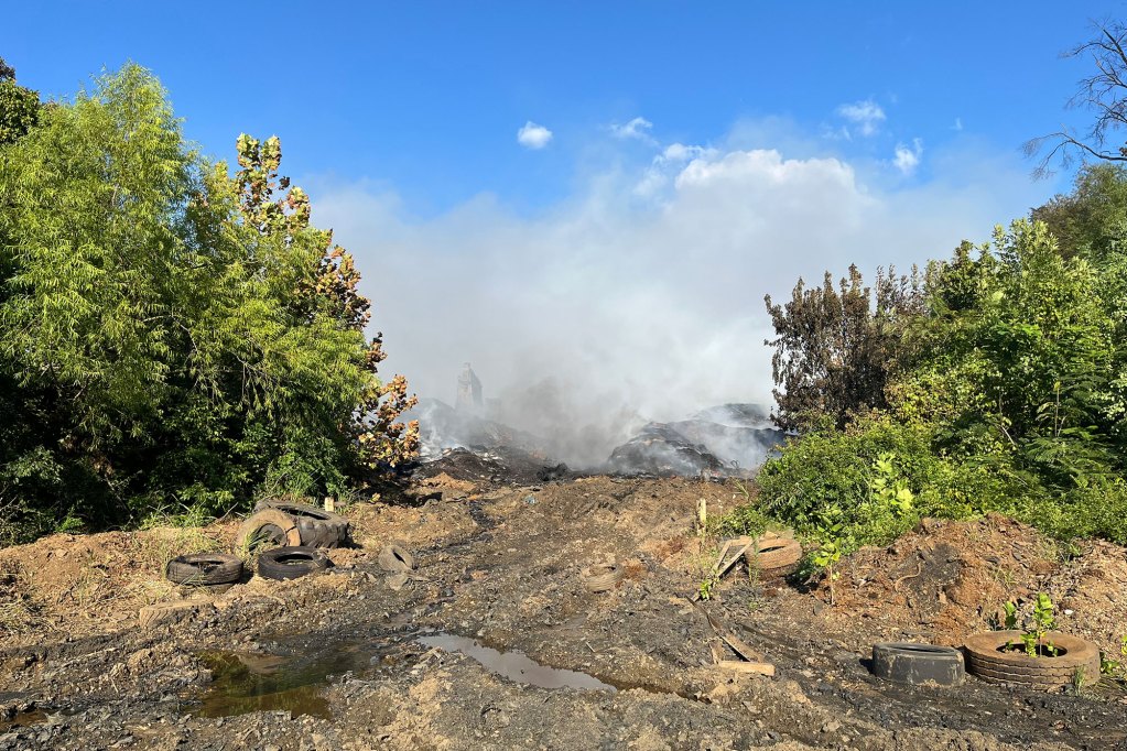Tire fire at a tire plant framed by trees on either side. Tires are visible in the dirt below the smoke