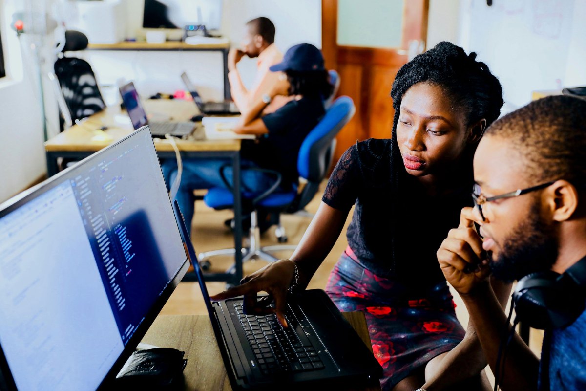 A man and a woman look at a computer screen together at a desk in a large room. Another desk with more people can be seen in the background