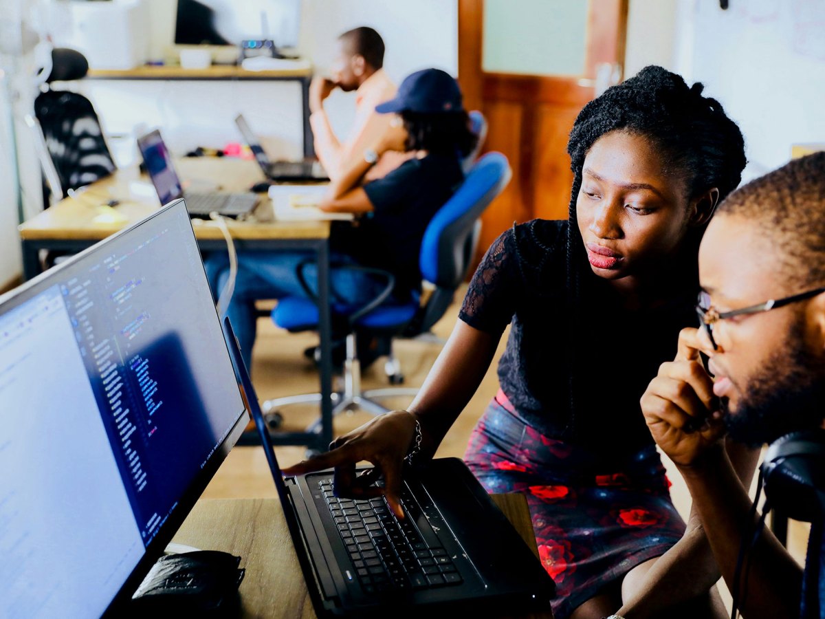A man and a woman look at a computer screen together at a desk in a large room. Another desk with more people can be seen in the background
