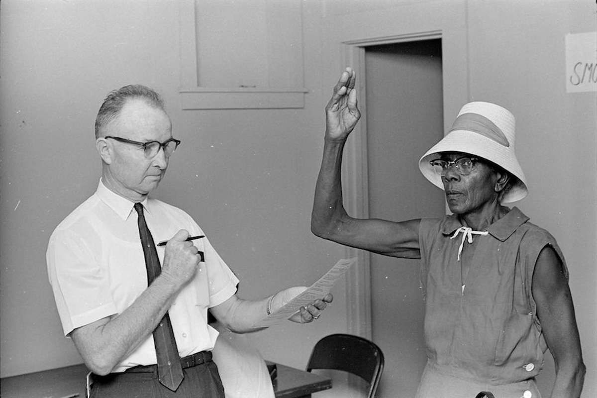 A black and white photo of a white man on the left with pen and paper in hand, and a black women on the right with one hand in the air