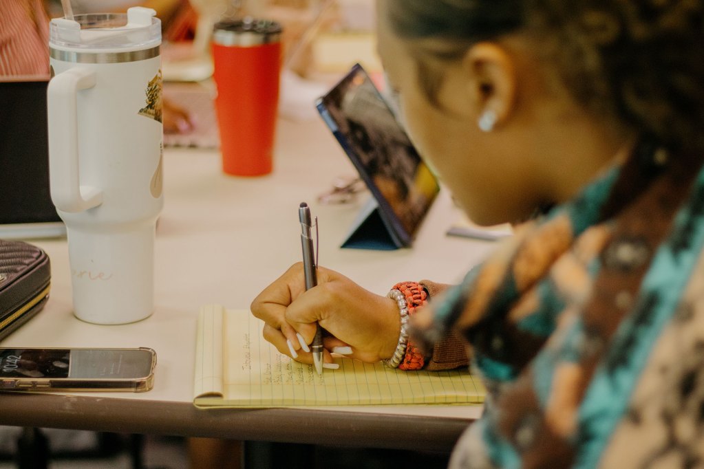 A closeup of a teen writing notes down on a yellow legal pad at a desk with other students also working