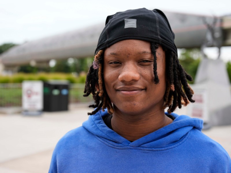 A teen in blue sweatshirt and black hat stands outside, smiling