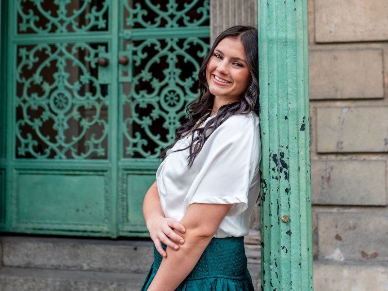 A teen leans against a green pole in front of a green door