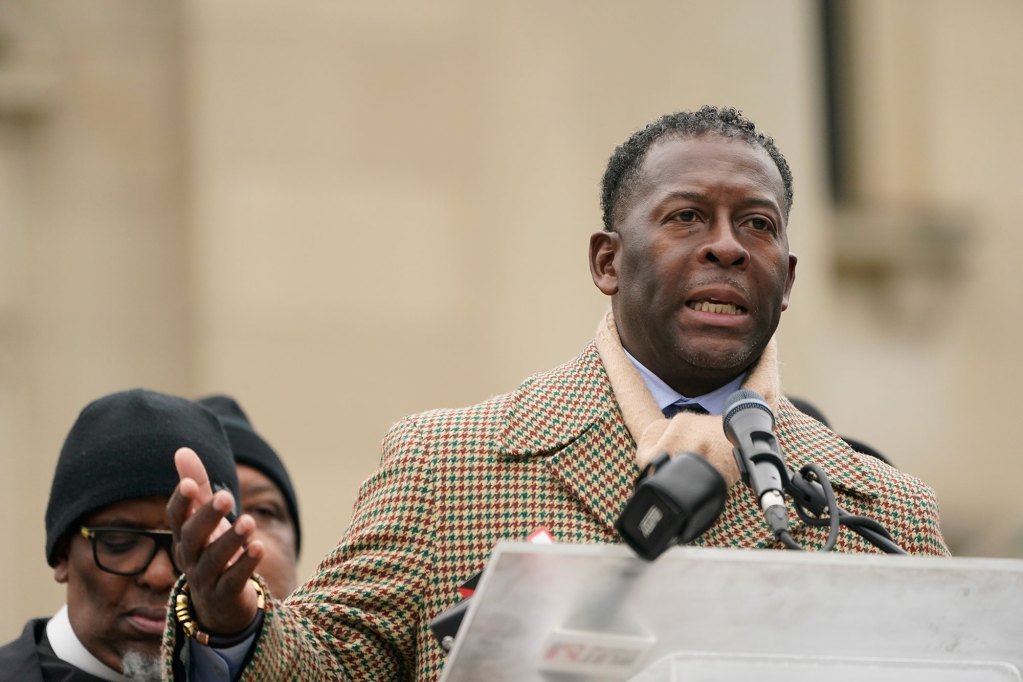 A man in a brown tone winter outfit speaks at a podium. Other men can be seen behind him