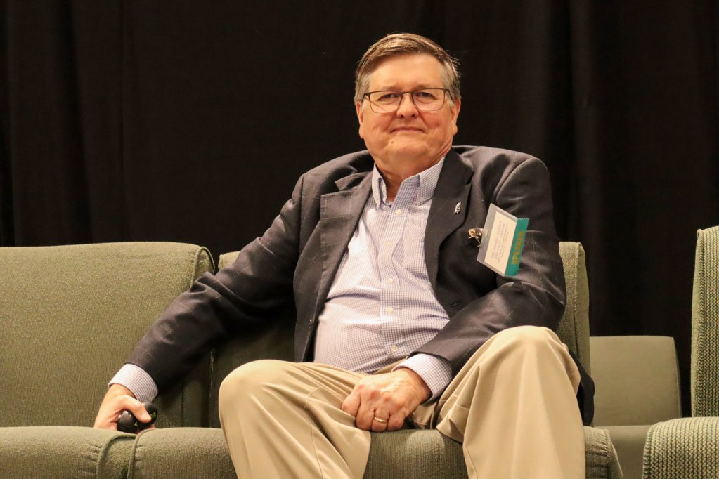 A man in a suit sits on a beige couch on a stage
