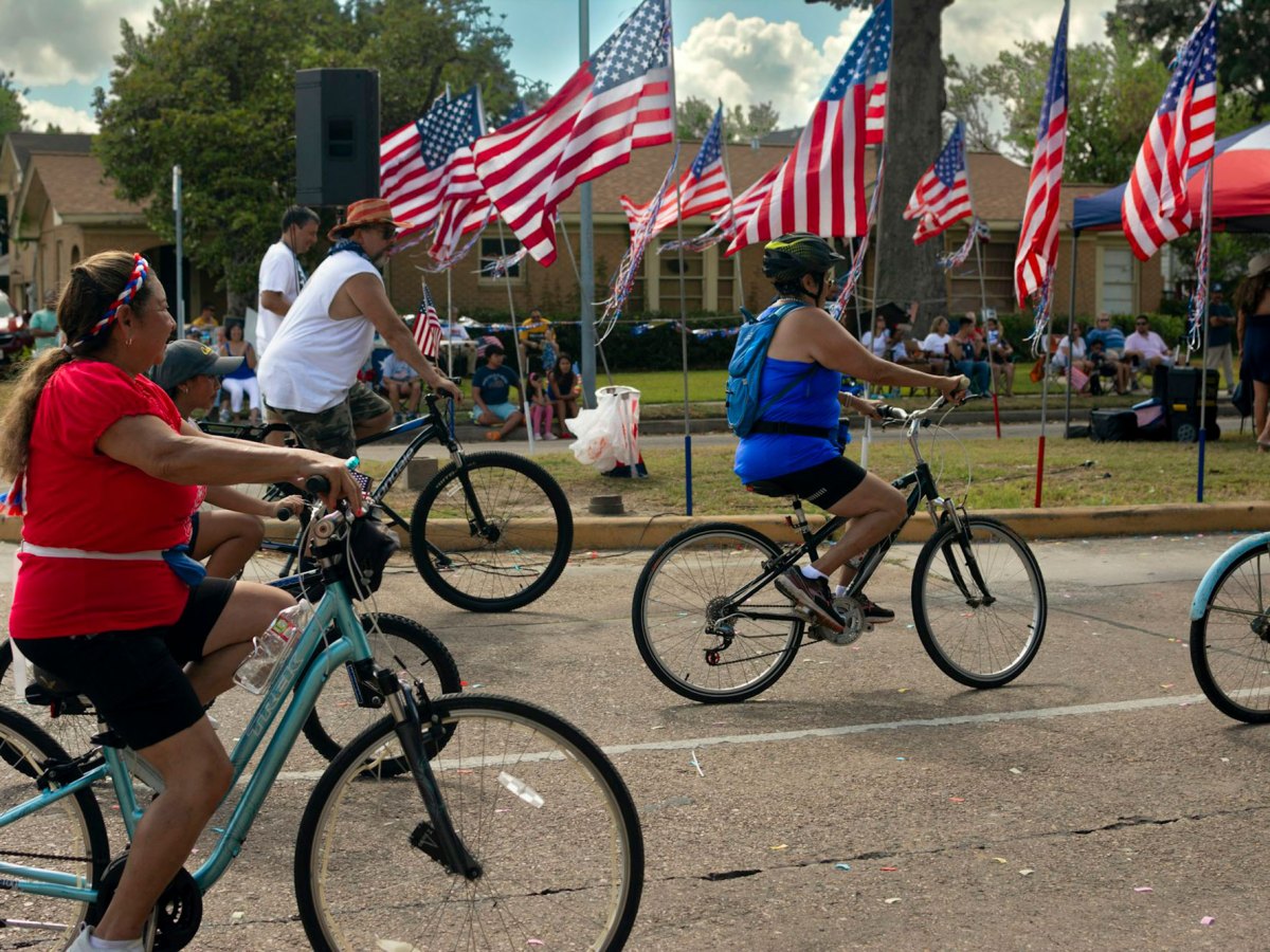 Families ride bicyles down the road past a road side decoration of multiple US Flags