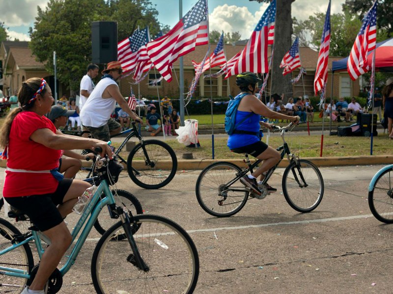 Families ride bicyles down the road past a road side decoration of multiple US Flags