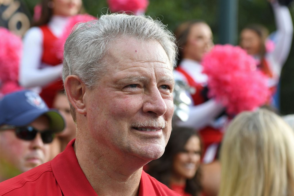Closeup of an older man in a red shirt, cheerleaders in red uniforms seen behind him in a crowd