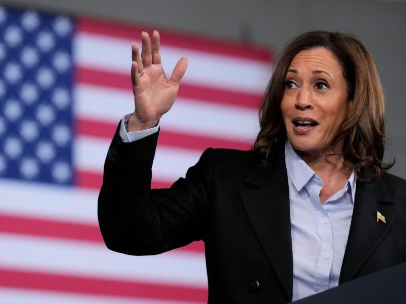 A woman in a black suit jacket and blue button up shirt waves while speaking. The US flag can be seen behind her