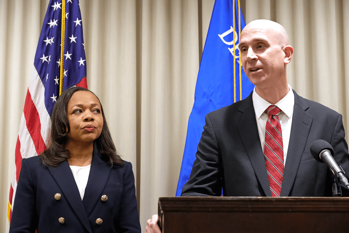 Two people stand before flags at a press conference. A bald man in a suit stands at a podium while a shorter woman in a suit looks on from the left.