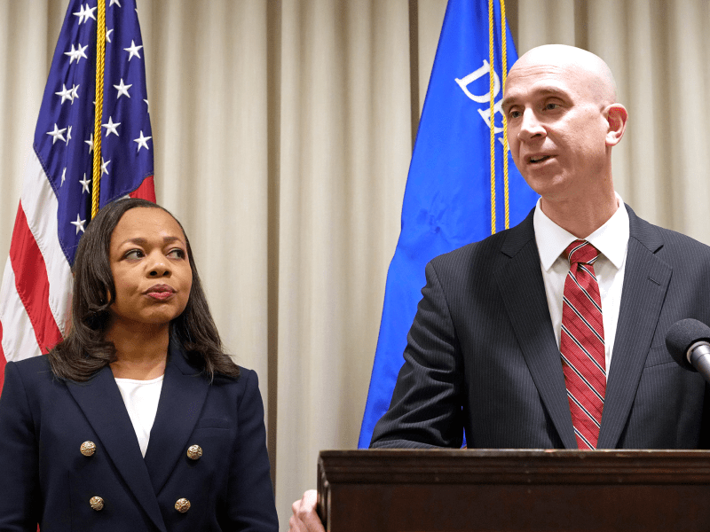 Two people stand before flags at a press conference. A bald man in a suit stands at a podium while a shorter woman in a suit looks on from the left.