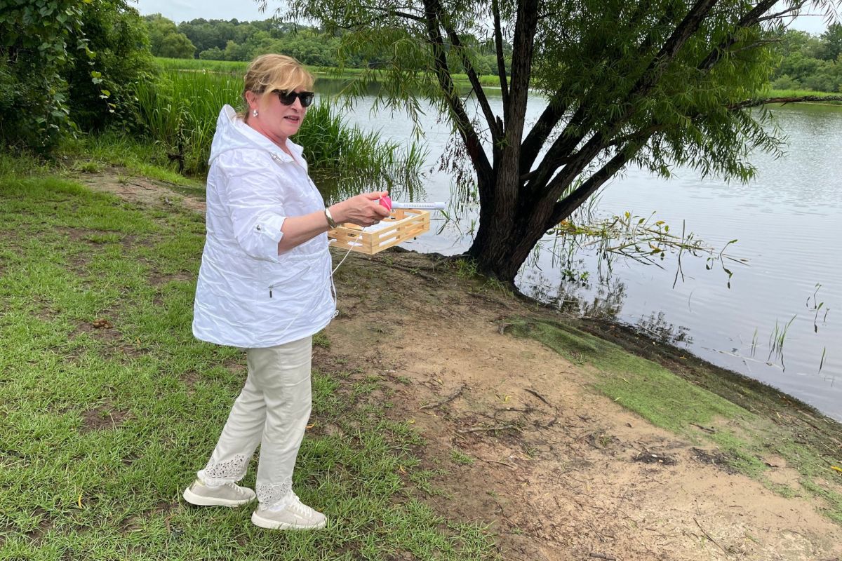 A woman in white stands beside A lake surrounded by green grass and trees, red vial in hand