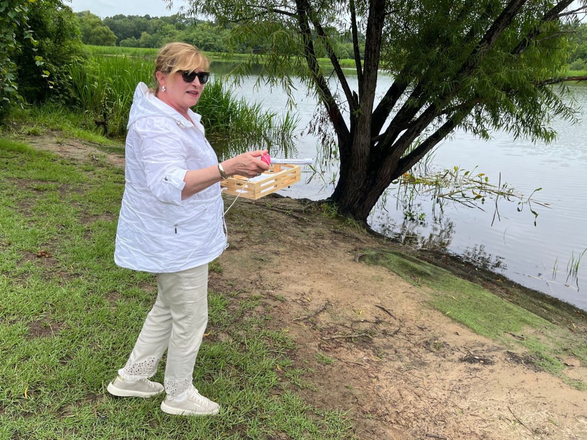 A woman in white stands beside A lake surrounded by green grass and trees, red vial in hand