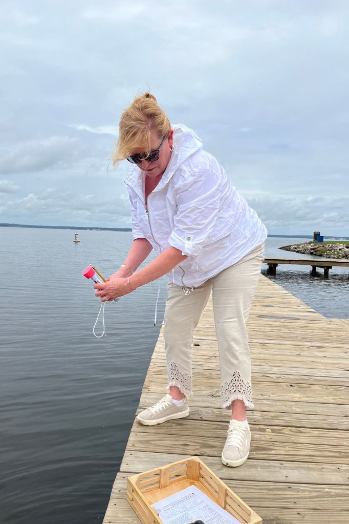 A woman stands on a wooden pier over the water with a red vial in her hand