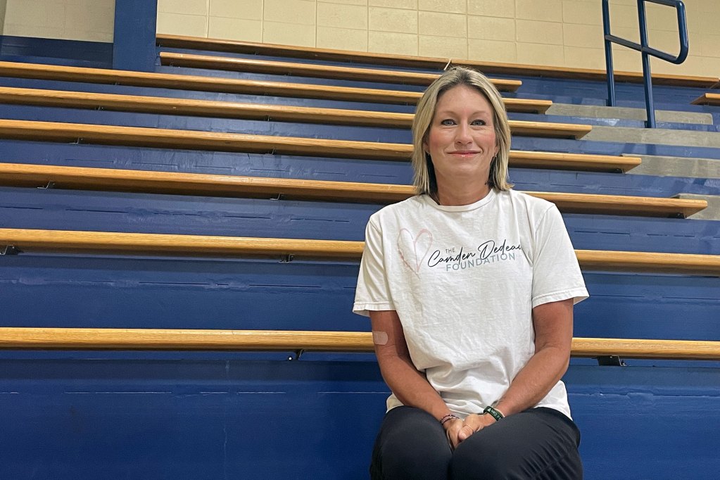 A smiling woman sits on blue painted sports bleachers. Her shirt reads The Camden Defense Foundation