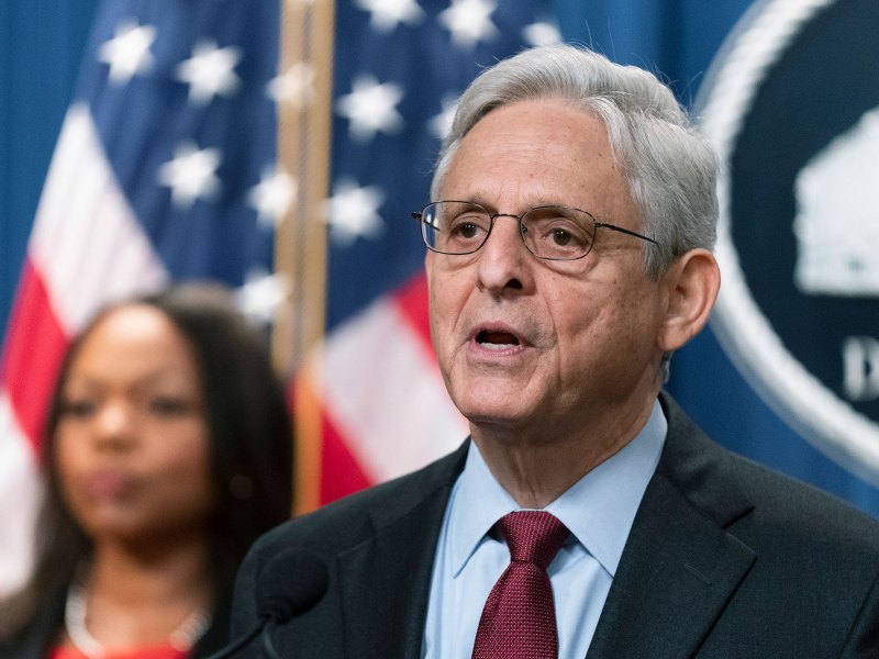 A man in a suit speaks. Out of focus behind him is a woman looking ahead standing in front of the US Flag