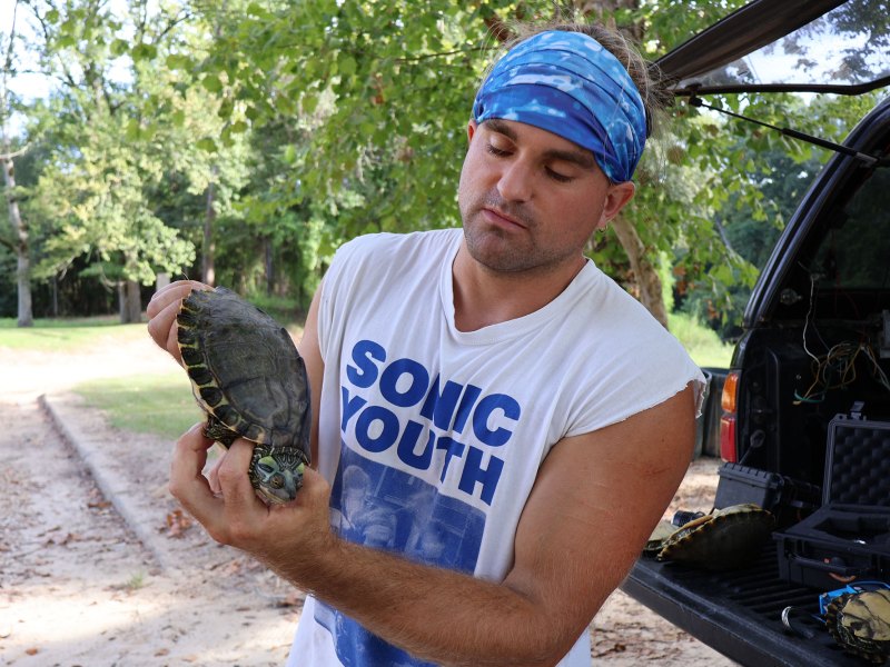 A man in a Sonic Youth tshirt and blue bandana holds a turtle behind his car