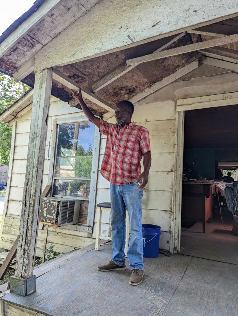 A man in jeans and a red patterned shirt stands on the porch of an older white building