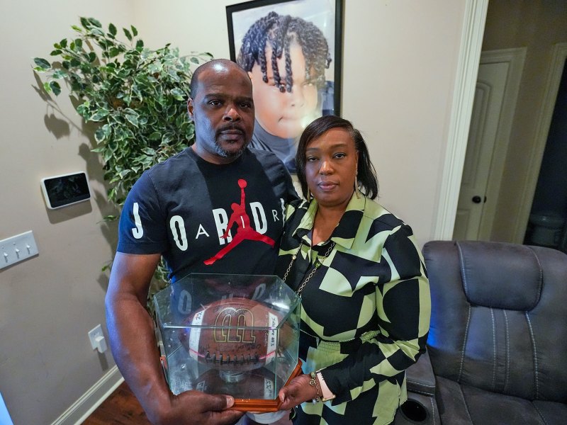 Phillip Laster Sr. and his wife Ashanta Laster stand in their living room and hold a football signed by teammates of their son Phillip Laster Jr.