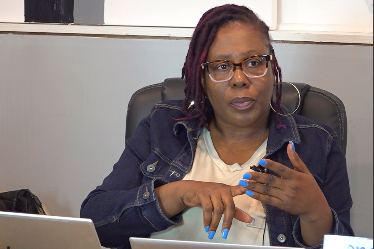 A seated woman in glasses speaks while gesturing with her hands. Her nails are painted light blue.