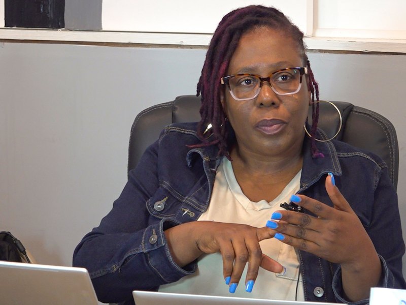 A seated woman in glasses speaks while gesturing with her hands. Her nails are painted light blue.