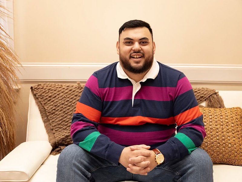 A man in a colorful striped shirt sits on a cream colored couch with brown accent pillows