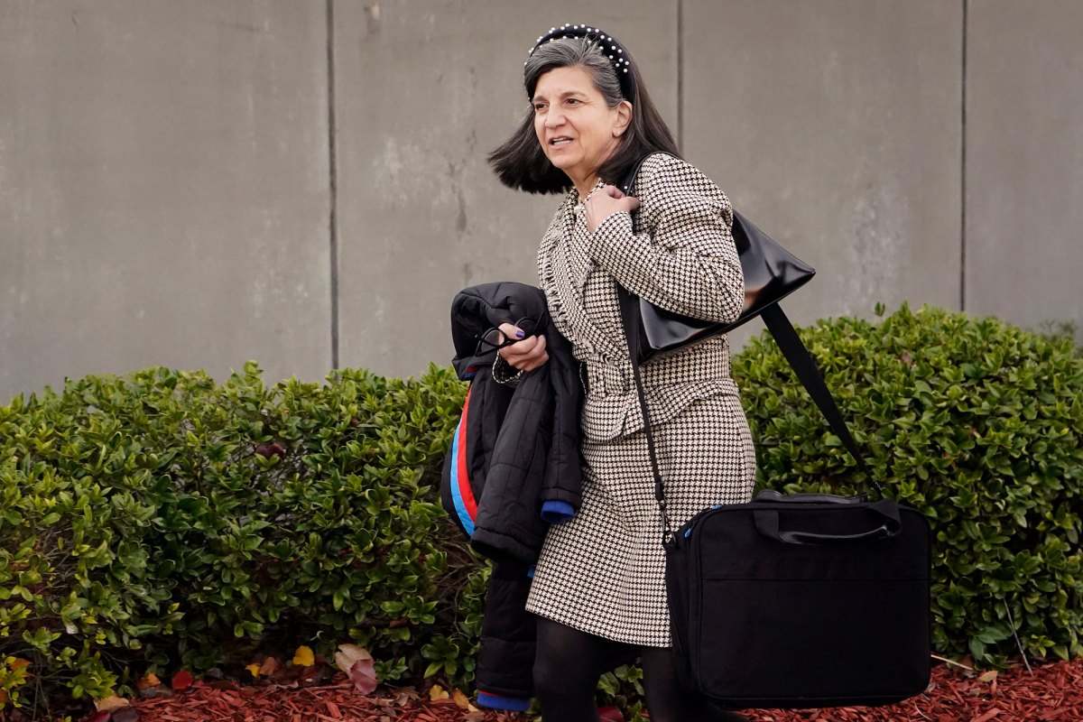A woman with black work bags hanging from her shoulder and a black coat under the other arm walks outside past a building