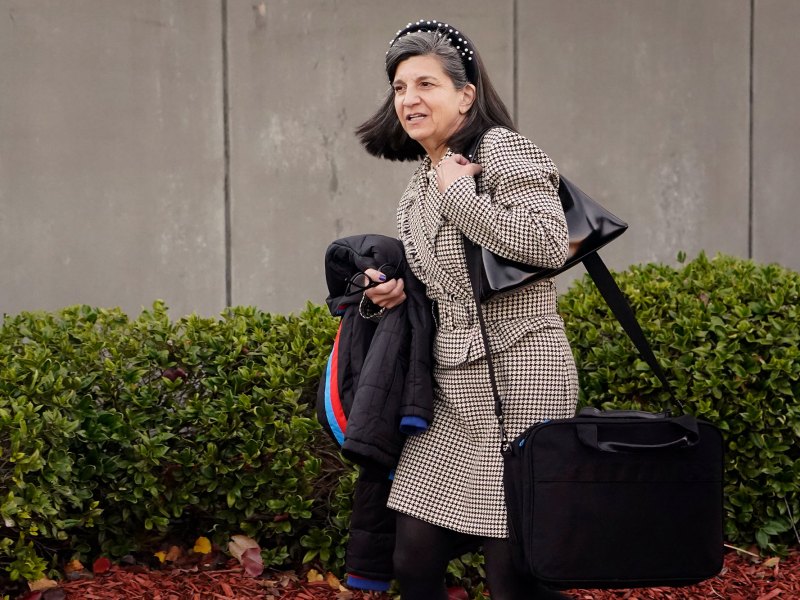 A woman with black work bags hanging from her shoulder and a black coat under the other arm walks outside past a building