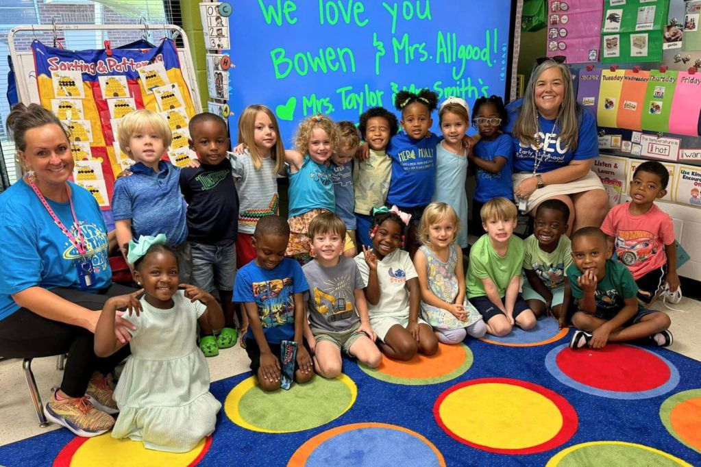 Two teachers sit in a colorful classroom with young students sitting and standing in two rows