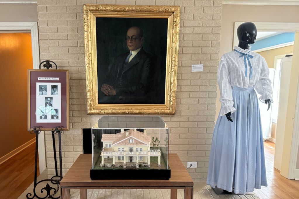An exhibit in the Utica Institute Museum showing a painting of a man, a model of a two story house, and a mannequin in a dress
