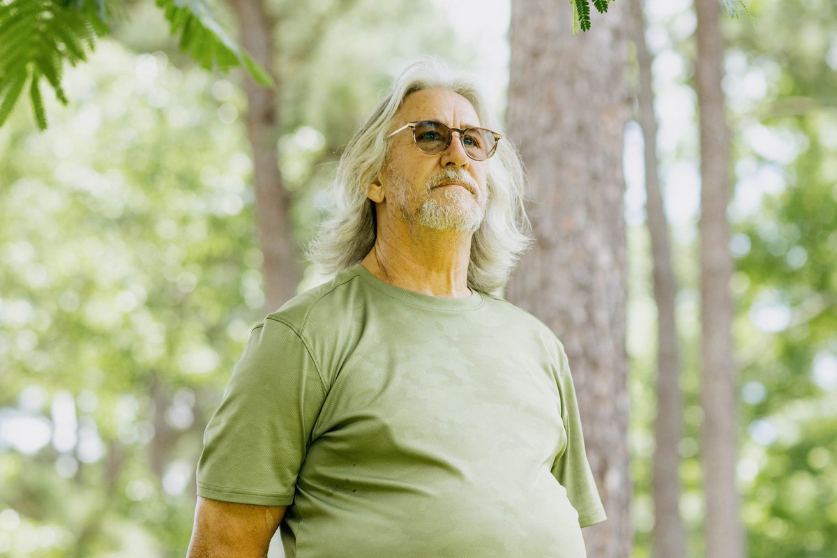 A photo of John Cook, a White man with gray shoulder-length hair, a gray beard, glasses and a green shirt. Cook is looking into the camera while standing outdoors under a tree.