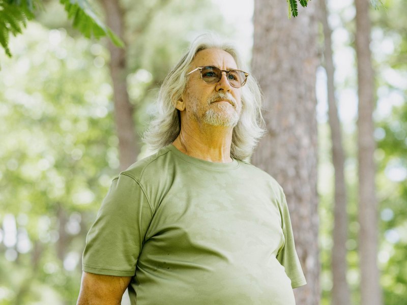 A photo of John Cook, a White man with gray shoulder-length hair, a gray beard, glasses and a green shirt. Cook is looking into the camera while standing outdoors under a tree.