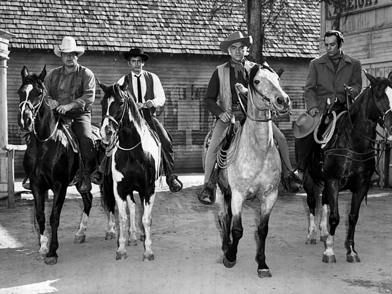 Four members of the cast of Bonanza seem riding horses through town