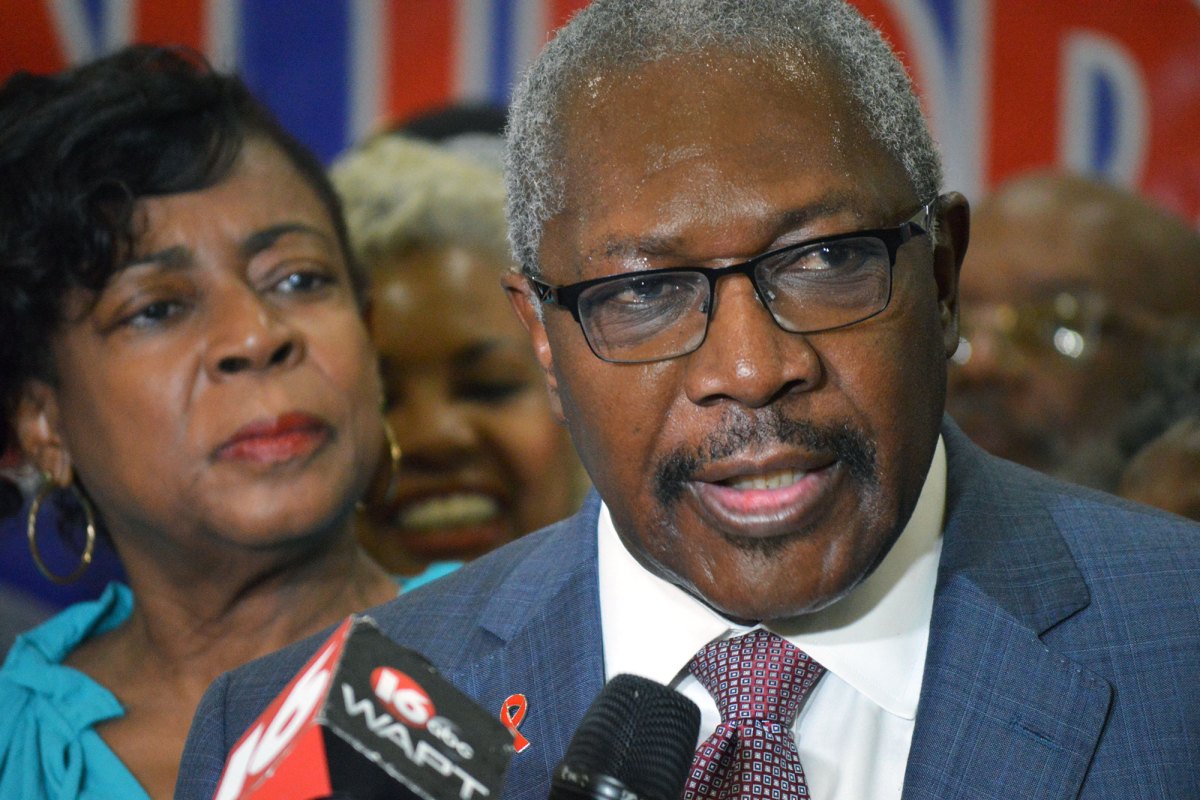 A man in glasses and a suit speaks int a reporter's mic as his wife and crowd look on behind him
