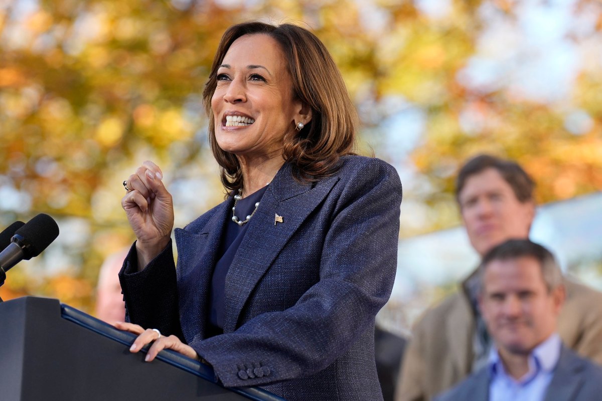 A woman speaks at a podium outside, the leaves in the trees have autumn colors